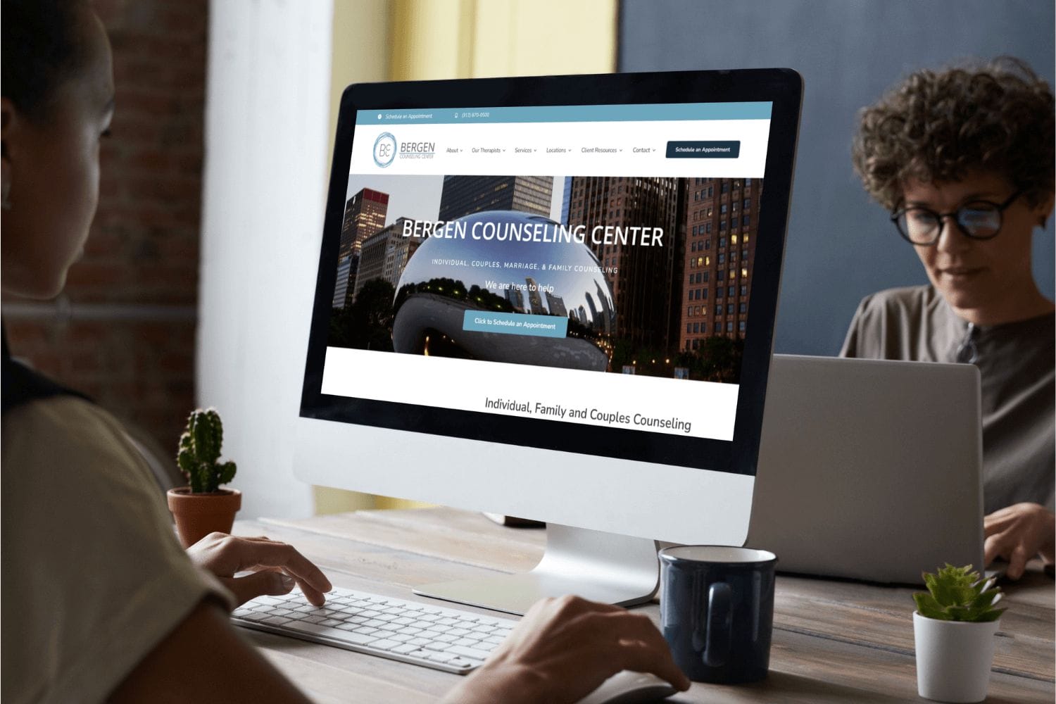 Two people are working at a desk with a computer displaying the Bergen Counseling Center's website, featuring options for scheduling appointments and information on individual, family, and couples counseling services in Chicago.