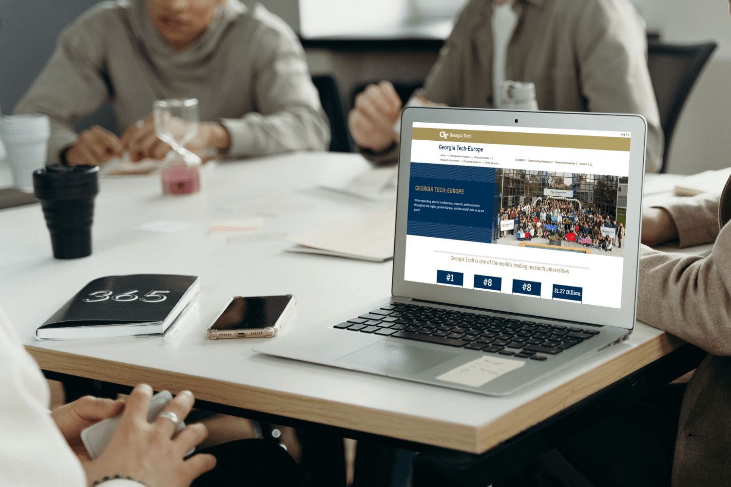 A group of people in a meeting sit around a table with notebooks and a laptop displaying a website, discussing the exciting rebrand of a top research university in Europe.
